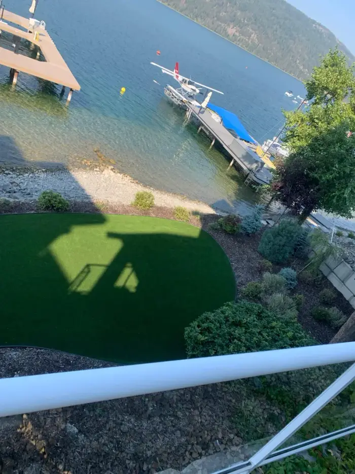 Elevated view of lush artificial turf installed at an Okanagan lakefront property with a seaplane moored at the dock and forested mountains reflected in the blue-green water.