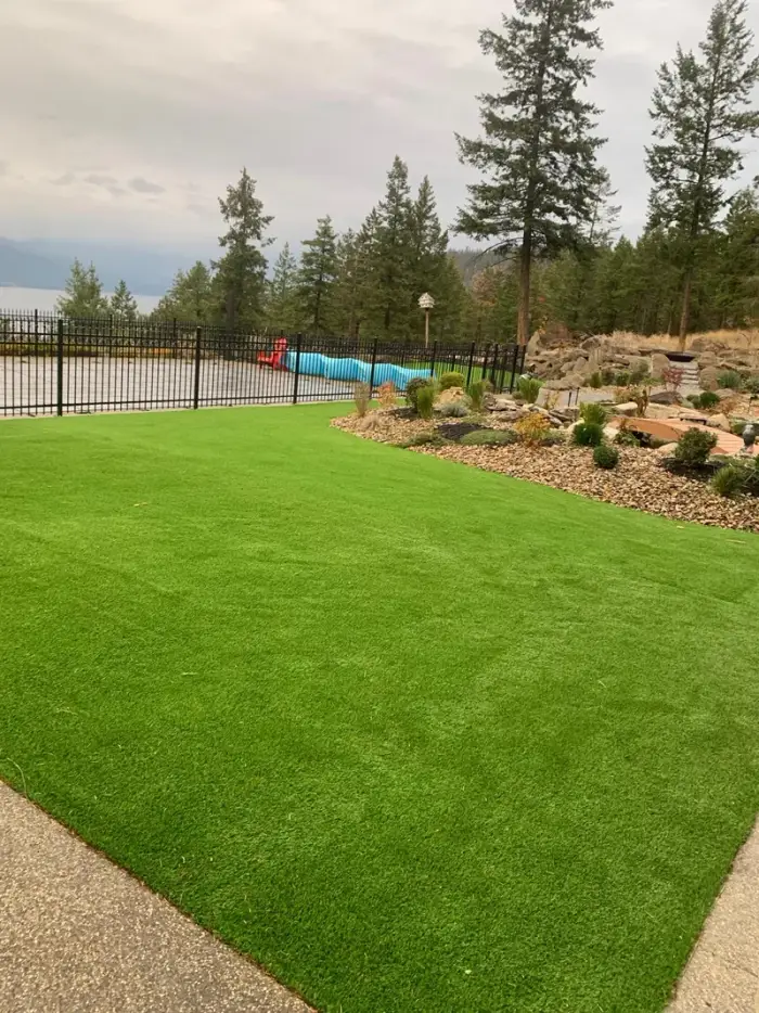 Vibrant artificial turf lawn with a river-rock garden border, black iron fence, and Okanagan Lake visible through the trees in the background.