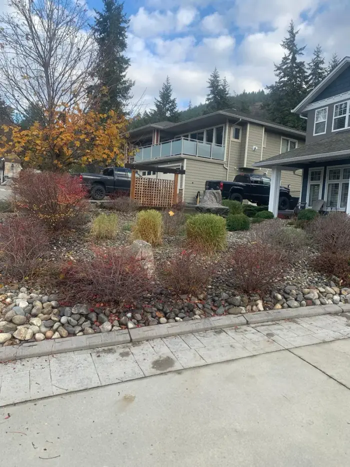 Established front yard rock garden with mixed ornamental shrubs in autumn red and yellow, rounded river rock edging, and a large feature boulder beside a residential Okanagan home.