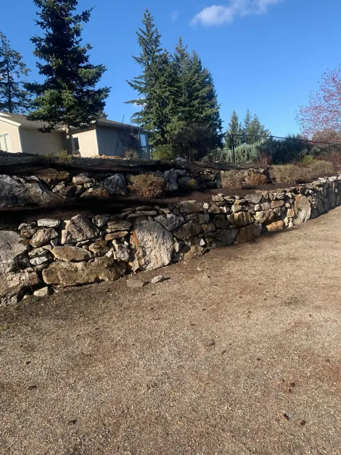 Dry-stacked natural stone retaining wall running across a rural Okanagan hillside property with pine trees behind and a dramatic mountain valley backdrop.