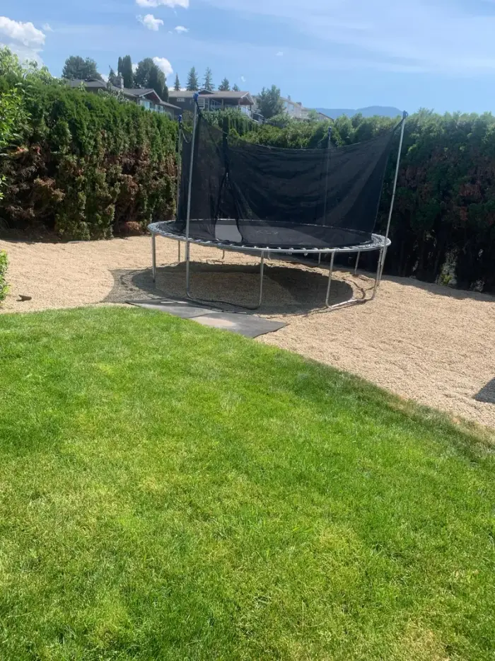 Freshly installed sod lawn with a trampoline area covered in pea gravel, surrounded by cedar hedges with Okanagan mountains visible over the rooftops.