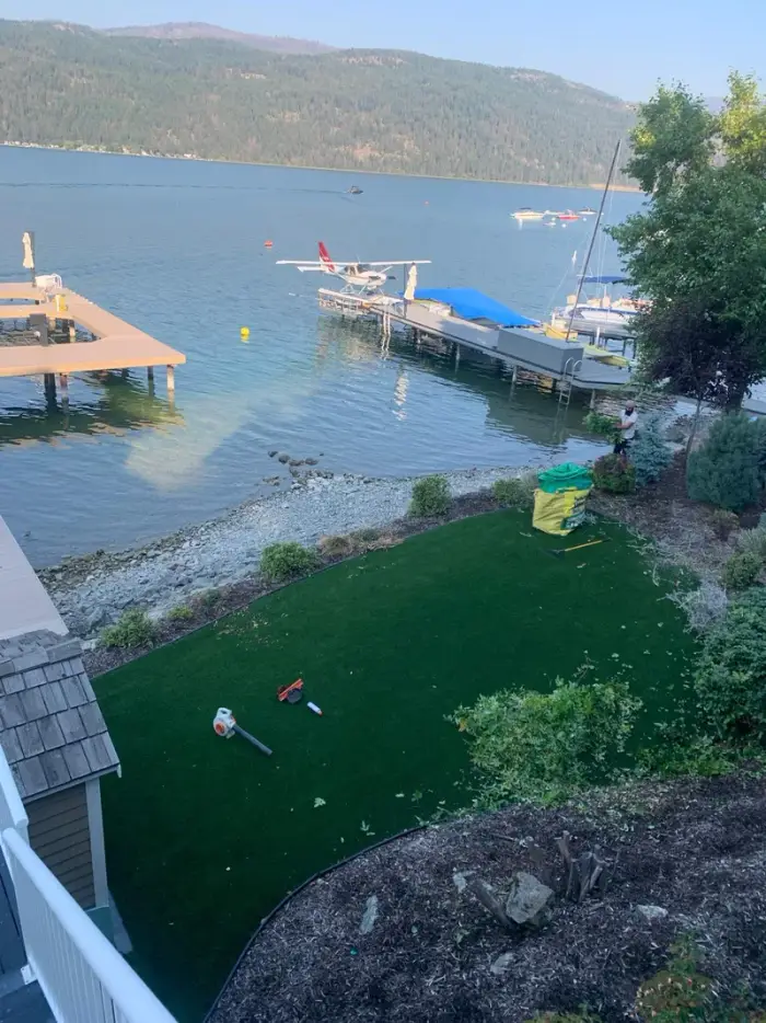 Crew member hand-weeding and tidying a front yard gravel landscape bed with large boulders and established shrubs at a residential Okanagan property.