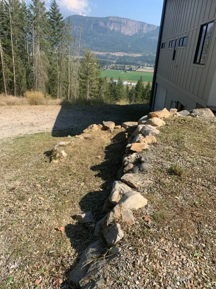 Boulder and field-stone edging line installed along a rural building foundation with a dramatic Okanagan mountain valley view stretching into the distance.