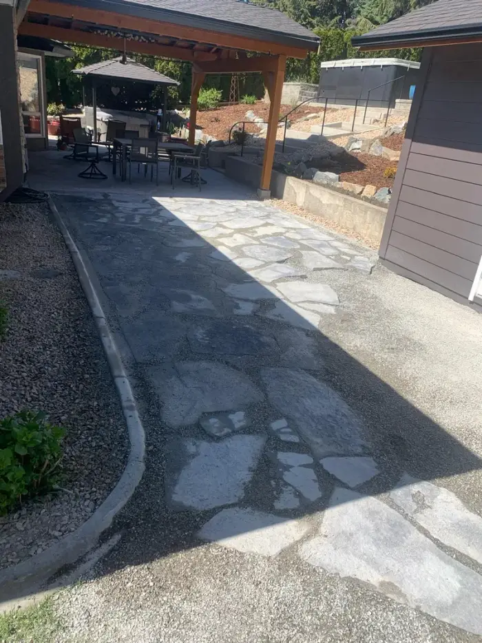 Large irregular flagstone patio recently installed beside a covered timber pergola with outdoor dining set, and freshly placed river rock mulch beds bordering the patio.