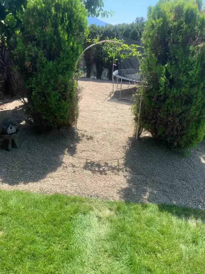Rustic garden archway made of natural branches framed by two trimmed cedar trees over a gravel path, with a manicured lawn and Okanagan mountain visible beyond.