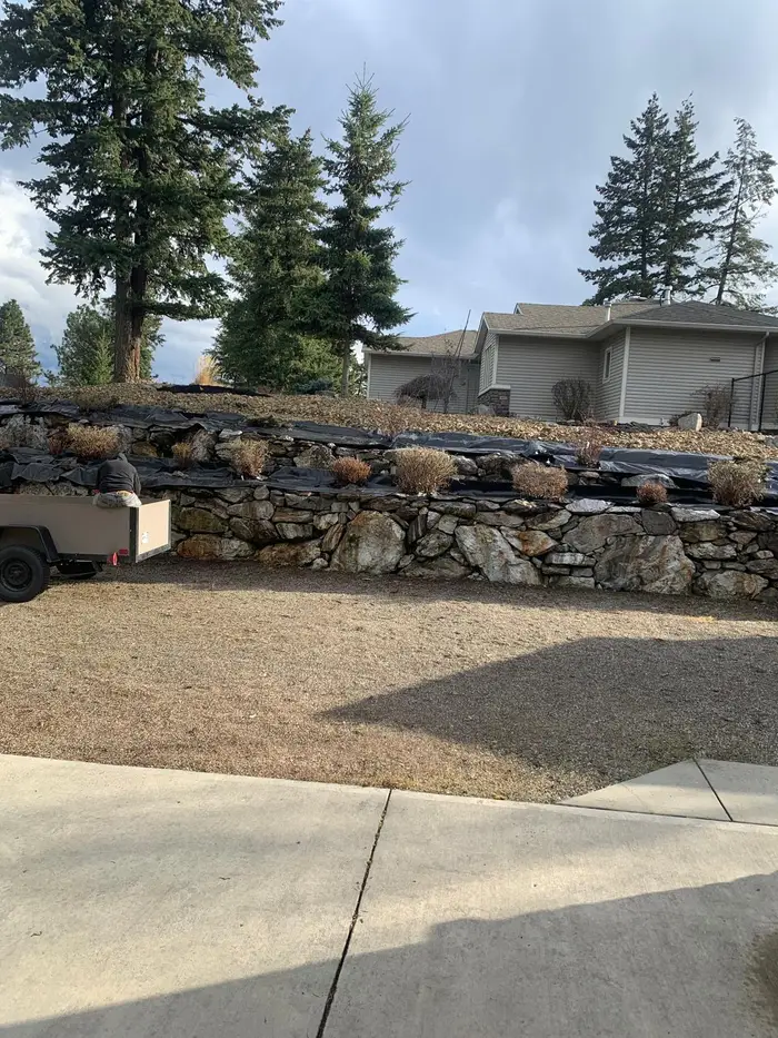 Double-tiered natural stone retaining wall with weed barrier and freshly planted low shrubs across the full width of a hillside residential property, trailer parked alongside.