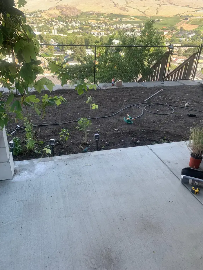 Freshly prepared hillside planting area with topsoil, drip irrigation installed, and new plantings going in — panoramic Okanagan city and valley view across rolling hills.