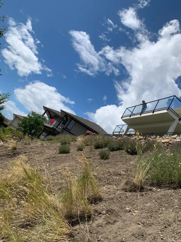 Crew member planting drought-tolerant grasses on a steep hillside below a modern Okanagan home with dramatic summer clouds overhead.