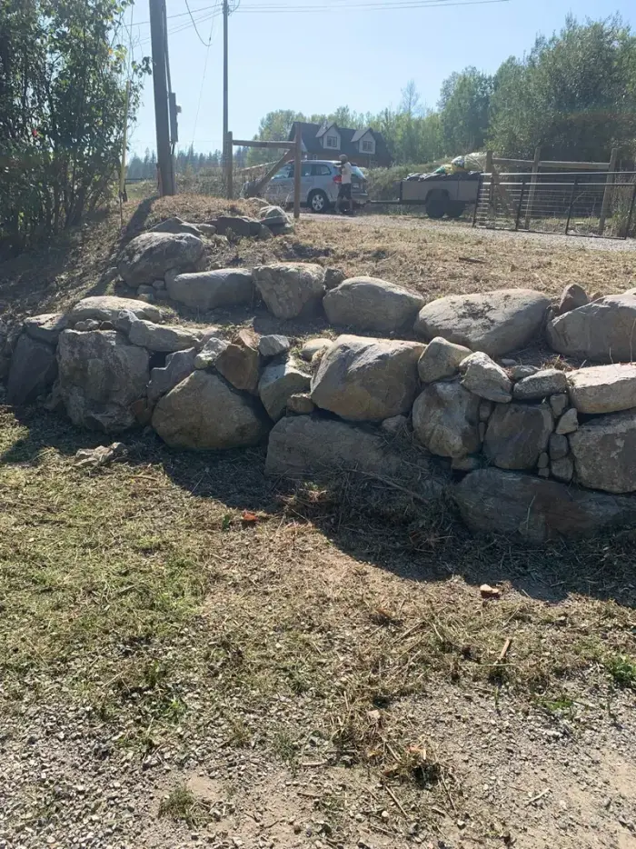 Natural boulder and field-stone retaining wall under construction in an open rural area, with cool Okanagan mountain and sky backdrop.