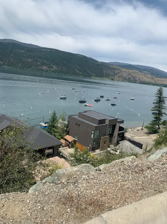 Dramatic aerial view looking down a rocky hillside at a modern lakefront home with dozens of boats anchored in Okanagan Lake and forested mountains across the water.