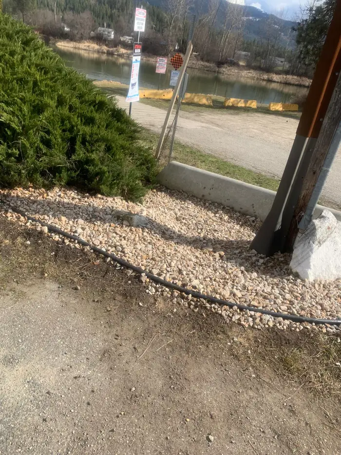White crushed-rock gravel edging installed beside a juniper hedge along a roadside near a river, with drip irrigation line visible and mountains across the valley.