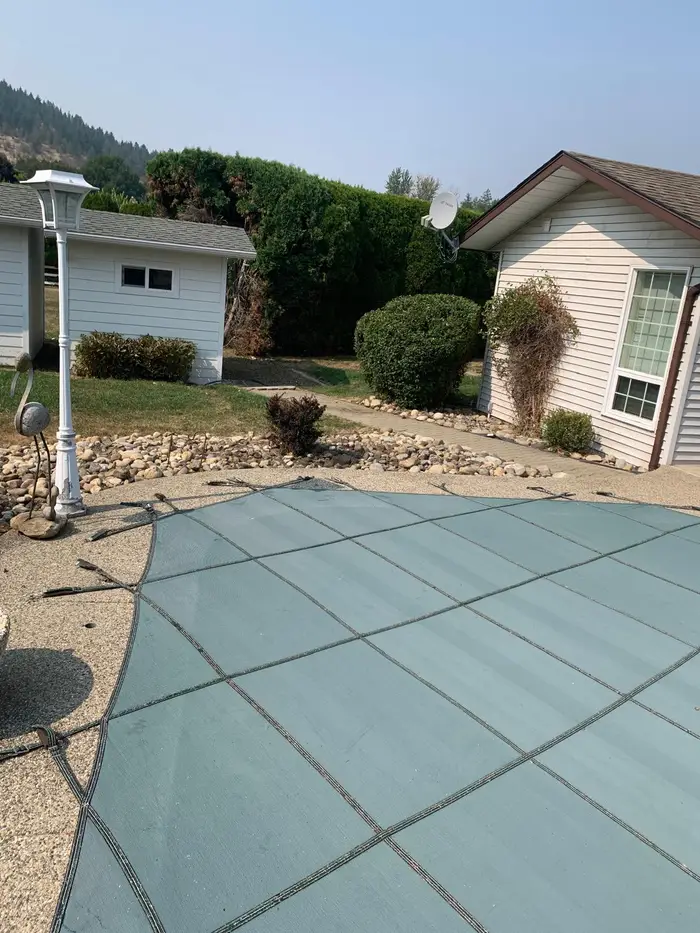 Backyard with a large teal pool safety cover, trimmed shrubs, river rock planting beds, and a maintained lawn area on a hazy summer day.